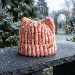 Può includere: Un berretto in maglia grossa in filato variegato color pesca e bianco, caratterizzato da due forme a punta di orecchie di gatto sulla parte superiore e un bordo ripiegato. Una piccola etichetta quadrata in pelle beige con icona di gatto.
