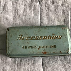 May include: A rectangular, light blue metal storage tin with rounded corners. The lid features the embossed text "Accessories" in a stylised script font above the words "SEWING MACHINE" in block letters. The tin shows signs of rust and wear.