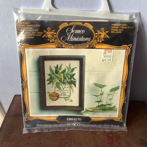 Puede incluir: Un kit de bordado crewel Semco Miniatures en una bolsa de plástico transparente. El empaque muestra un diseño terminado de chiles verdes en un frasco y un tazón pequeño, con el texto Chilli 771 y un tamaño final de 127 mm x 178 mm.