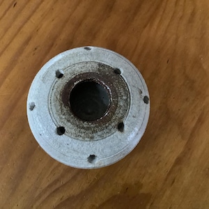 May include: A circular ceramic object with a textured, off-white exterior and a dark, weathered brown centre. The piece features a central opening surrounded by six evenly spaced small holes arranged in a ring pattern on the flat top surface.