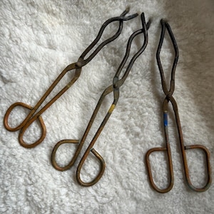 May include: Three vintage metal crucible tongs with scissor-style handles and curved gripping ends. The tongs feature a weathered, brownish-metallic finish with small patches of yellow and blue paint markings near the central pivot points.
