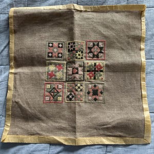 May include: A square piece of tan linen fabric featuring a central grid of nine embroidered cross-stitch squares. Each square contains a unique geometric pattern in shades of pink, green, yellow, and black. The fabric has a light yellow border.