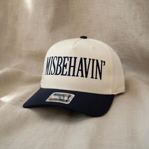May include: A cream-colored baseball cap with a navy blue brim. The text "MISBEHAVIN'" is embroidered in navy blue across the front. The cap has a curved brim and a small button on top.