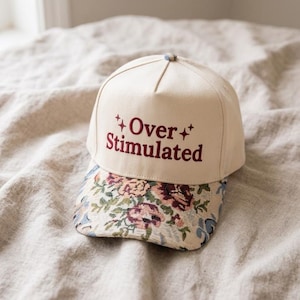 May include: A cream-colored baseball cap featuring the embroidered text Over Stimulated in dark red with small star accents. The brim of the cap is decorated with a detailed floral tapestry pattern in shades of pink, green, and blue.