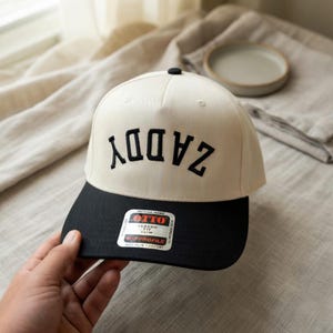 May include: A two-tone baseball cap featuring an off-white crown and a black brim. The word ZADDY is embroidered in black, arched, and upside-down across the front panels. A rectangular sticker on the brim reads OTTO CLASSIC FIT.