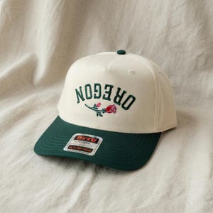May include: A cream-colored baseball cap with a dark green brim. The front features the word OREGON embroidered in dark green capital letters with an arched design, accompanied by a small, detailed red rose with green leaves centered below.