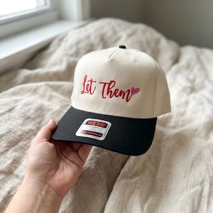 May include: A cream-colored baseball cap with a black brim. The phrase "Let Them" is embroidered in red script on the front, with a small red heart. The cap has a classic, structured design.