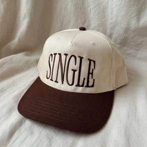 May include: A beige and brown baseball cap with the word "SINGLE" embroidered in dark brown letters across the front. The cap has a curved brim and a button on top.