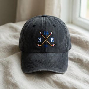 May include: A charcoal gray baseball cap with an embroidered hockey-themed design. The design features crossed hockey sticks, the letters "H" and "R", and the numbers "24" and "81" in blue, red, and yellow.