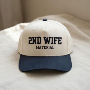 May include: A baseball cap with a cream-colored crown and a navy blue brim. The cap features the embroidered text "2ND WIFE" above "MATERIAL" in navy blue block letters. A navy blue button is on top.