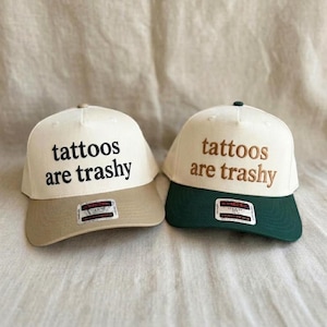 May include: Two baseball caps with off-white crowns and embroidered text reading tattoos are trashy. The left cap has a tan brim and black text, while the right cap features a dark green brim and brown text. Both include a small rectangular sticker.