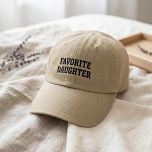 Może przedstawiać: Beżowa czapka z daszkiem z wyhaftowanym czarnym napisem "FAVORITE DAUGHTER". Czapka wykonana jest z miękkiej tkaniny i ma zakrzywiony daszek. Kapelusz leży na beżowej tkaninie.