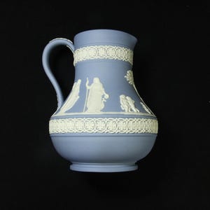 May include: A light blue ceramic pitcher with a curved handle. The pitcher features white relief designs of figures and decorative bands around the top and base. The background is black.