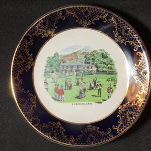 May include: Decorative plate with a blue border and gold filigree design. The center features a colorful illustration of a gathering in front of a building, with the word "LLANGOLLEN" at the bottom.