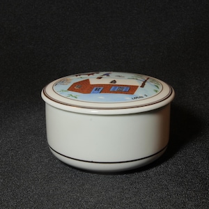 May include: A white ceramic trinket box with a lid featuring a painted scene of a house with a red roof, blue windows, and a chimney. The lid has a brown border and the box has a black trim.