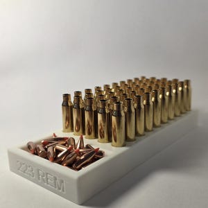 May include: A white tray containing numerous brass-coloured bullets with red tips, labelled "223 REM." The bullets are arranged in rows, with some loose in a separate compartment. The background is plain white.