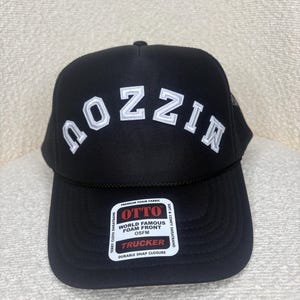 May include: Black trucker hat with the word "NOZZIN" in white block letters across the front. The hat has a mesh back and a curved brim. A white tag on the front reads "OTTO WORLD FAMOUS FOAM FRONT TRUCKER".