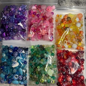 Könnte beinhalten: Sechs transparente Plastiktüten gefüllt mit bunten Perlen. Die Tüten enthalten Perlen in Lila, Rosa, Gelb, Blau, Grün und Rot. Die Perlen variieren in Größe und Form, darunter Sterne, Blumen und runde Perlen, ideal für die Schmuckherstellung.
