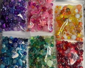 May include: Six clear plastic bags filled with colorful beads. The bags contain beads in shades of purple, pink, yellow, blue, green, and red. The beads vary in size and shape, including stars, flowers, and round beads, perfect for jewelry making.