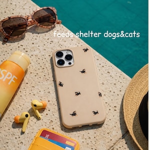 Könnte beinhalten: Eine beige Handyhülle mit einem Muster aus kleinen schwarzen Hundeillustrationen. Das Bild zeigt auch Sonnenbrillen, Sonnencreme, gelbe Ohrhörer, ein gelbes Portemonnaie und einen Strohhut. Der Text "feeds shelter dogs&cats" ist sichtbar.