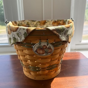 May include: A round woven wooden basket featuring a fabric liner with a pumpkin and leaf pattern. The basket includes a decorative oval tag on the front displaying a printed illustration of three pumpkins and green leaves, accented by green woven strips.