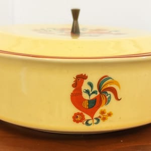 May include: A pale yellow ceramic casserole dish with a matching lid and a dark brown knob handle. The dish features a colorful rooster graphic in red, blue, and orange, with integrated side handles and a thin brown rim accenting the lid edge.