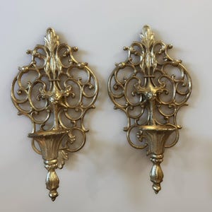 May include: A pair of gold-coloured metal wall sconces featuring intricate scrollwork and filigree patterns. Each sconce has a central vertical spine topped with a fleur-de-lis motif, a small cup-shaped candle holder, and a tapered finial at the base.