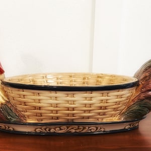 May include: A ceramic serving bowl shaped like a rooster with a basket-weave pattern in the center. The rooster has a red comb and wattle with cream and brown feathers. The tail feathers are dark brown and green. The base has a black trim.