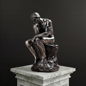 Könnte beinhalten: Eine metallisch silberfarbene Skulptur einer sitzenden Figur in kontemplativer Haltung, die ihr Kinn auf eine Hand stützt. Die Figur sitzt auf einem strukturierten, felsartigen Sockel, der auf einem weißen Marmorpodest mit grauer Maserung steht.