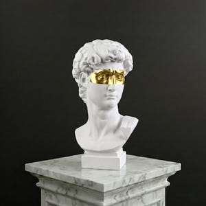 May include: A white bust of David with a gold-coloured mask covering the eyes. The sculpture is on a white marble pedestal against a dark background. The bust is a classical art piece.