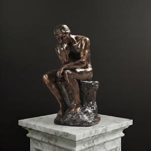 Pode incluir: Uma escultura em tom de bronze de uma figura sentada em pose contemplativa, com o queixo apoiado em uma das mãos. A figura está sobre uma base texturizada semelhante a uma rocha, exibindo musculatura detalhada e um acabamento metálico.