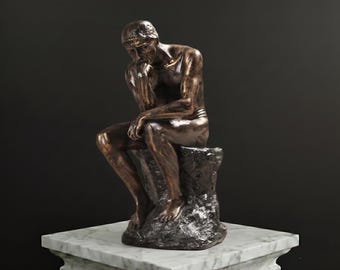 May include: A bronze-toned sculpture of a seated figure in a contemplative pose, resting the chin on one hand. The figure is perched on a textured, rock-like base, showcasing detailed musculature and a metallic finish with dark highlights.