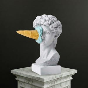 May include: A white sculpture of a classical head with an ice cream cone through the face. The cone is yellow and the ice cream is light blue. The sculpture is on a white marble plinth.