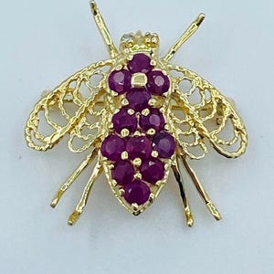 Puede incluir: Broche de abeja dorada con piedras preciosas de rubí. El broche presenta un diseño de filigrana detallado para las alas y las patas, con un grupo de rubíes redondos que forman el cuerpo. La abeja tiene una pequeña corona en la cabeza.