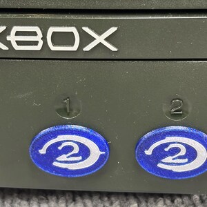 May include: A dark gray Xbox console front panel featuring the silver Xbox logo at the top. Below the logo, two circular blue stickers with a white number 2 graphic are positioned over the controller ports, which are marked with numbers 1 and 2.