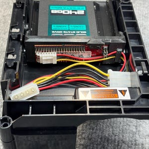 Puede incluir: Una carcasa de disco duro negra con una unidad de estado sólido (SSD) instalada. La SSD tiene una etiqueta turquesa con texto blanco que indica su capacidad y velocidad. El cableado y los conectores son visibles, junto con las etiquetas de advertencia.