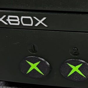 May include: A black Xbox video game console featuring the silver Xbox logo text on the front panel. Below the logo are two circular controller ports labeled with the numbers one and two, each displaying a bright green X symbol in the center.
