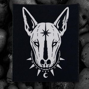 May include: Black square patch featuring a white illustration of a dog's head with a star on its forehead and a spiked collar with a crescent moon. The dog has pointed ears and a stern expression. The patch is set against a background of skulls.