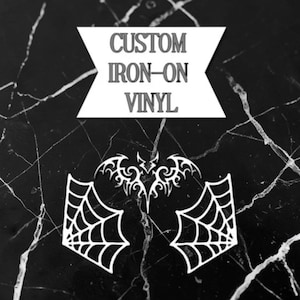 May include: White custom iron-on vinyl designs on a black marble background. The designs include a bat, spiderwebs, and the text "CUSTOM IRON-ON VINYL" in a banner.