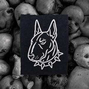 Puede incluir: Parche negro rectangular con un contorno blanco de la cabeza de un perro Bull Terrier y un collar de púas. El fondo es una colección de cabezas de muñecas antiguas monocromáticas.