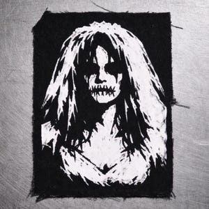 May include: Black and white patch featuring a stylized illustration of a woman's face with long hair and a stitched-up mouth. The design is a stark contrast against the black fabric.