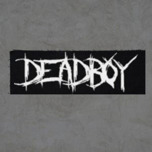 May include: A black rectangular patch with the white text "DEADBOY" in a distressed, gothic font. The patch has a rough, frayed edge, and the background is a neutral gray.