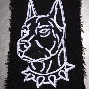 Puede incluir: Parche de tela negra con un contorno blanco de la cabeza de un perro de perfil. El perro tiene orejas puntiagudas y un collar de púas. Los bordes del parche están deshilachados, lo que le da un aspecto crudo e inacabado. El diseño es audaz y gráfico.