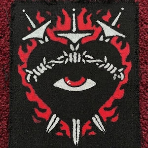 May include: Black patch featuring a heart-shaped design with a central eye, surrounded by barbed wire and flames. White swords and flames accent the design. The patch is likely for clothing or accessories.