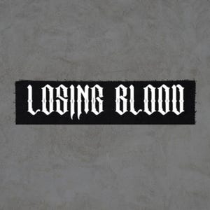 May include: A black rectangular patch with the words "LOSING BLOOD" in white gothic lettering. The patch has a frayed edge and is set against a gray background.