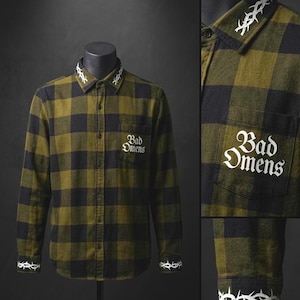 May include: A long-sleeved, olive green and black plaid shirt with white embroidered barbed wire details on the collar and cuffs. The words "Bad Omens" are embroidered on the chest pocket in a gothic font. The shirt is displayed on a black mannequin.