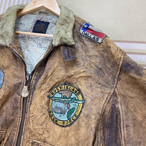 May include: A vintage brown leather jacket with a distressed finish, featuring a fur-lined collar and multiple patches. Patches include "DALLAS" and "BUSY ISLAND TASK FORCE". The jacket is hanging on a wooden hanger.