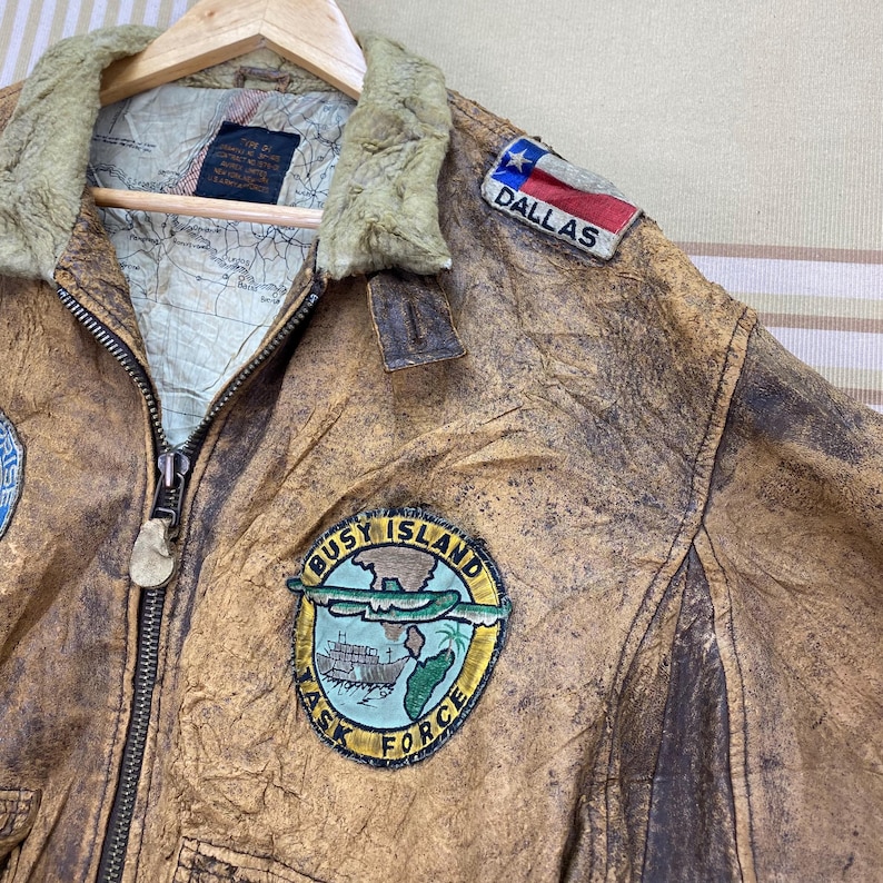 May include: A vintage brown leather jacket with a distressed finish, featuring a fur-lined collar and multiple patches. Patches include "DALLAS" and "BUSY ISLAND TASK FORCE". The jacket is hanging on a wooden hanger.