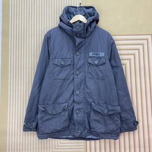 May include: A blue-grey jacket with a hood, multiple pockets, and button closures. The jacket features the brand name "AVIREX" on a small patch. The jacket has a casual, utilitarian design.