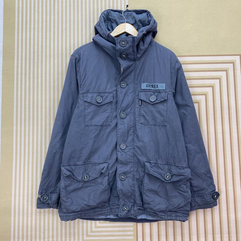 May include: A blue-grey jacket with a hood, multiple pockets, and button closures. The jacket features the brand name "AVIREX" on a small patch. The jacket has a casual, utilitarian design.